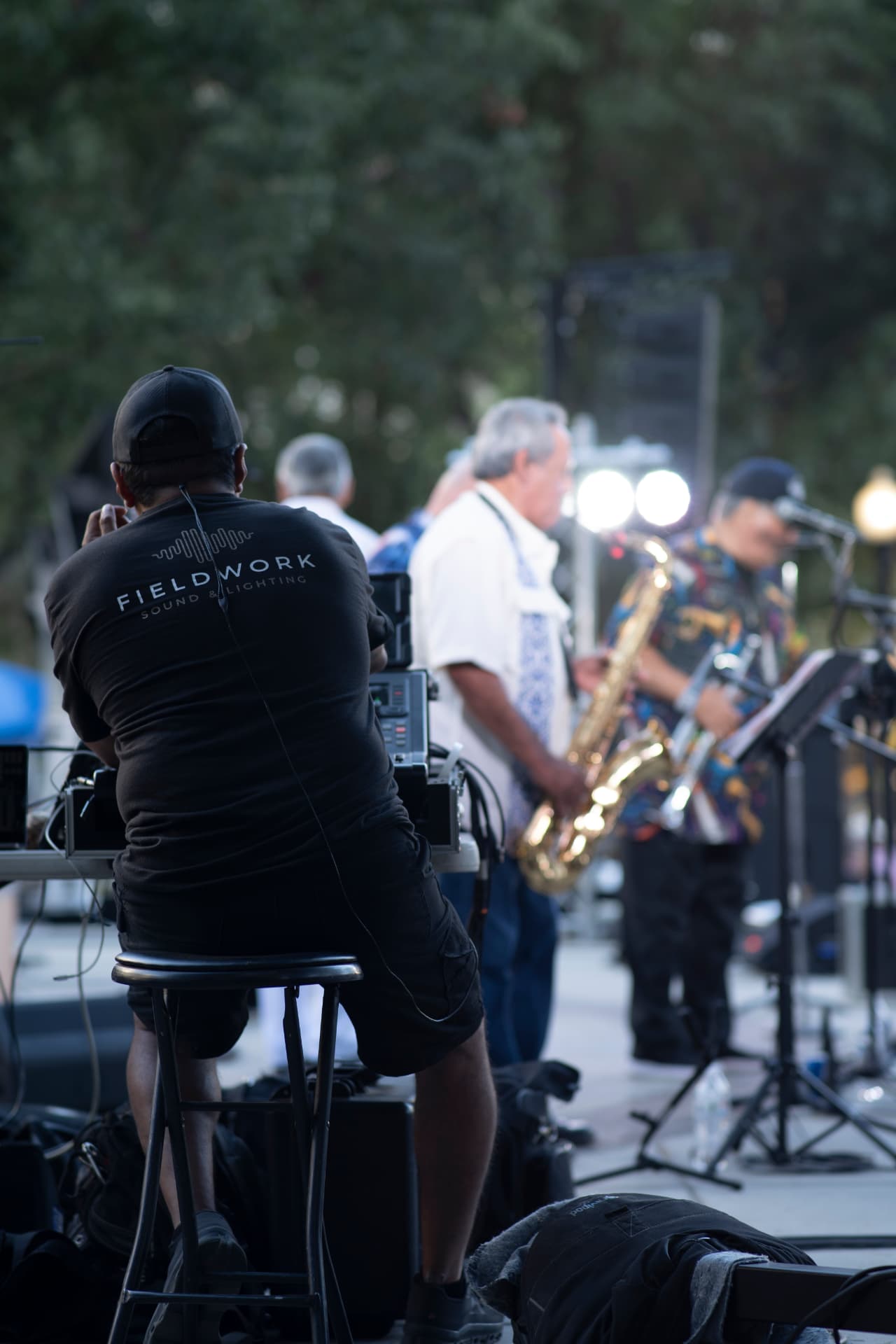 Sound technician in a Fieldwork Sound & Lighting shirt working a mixing board during an outdoor concert with a saxophone player on stage