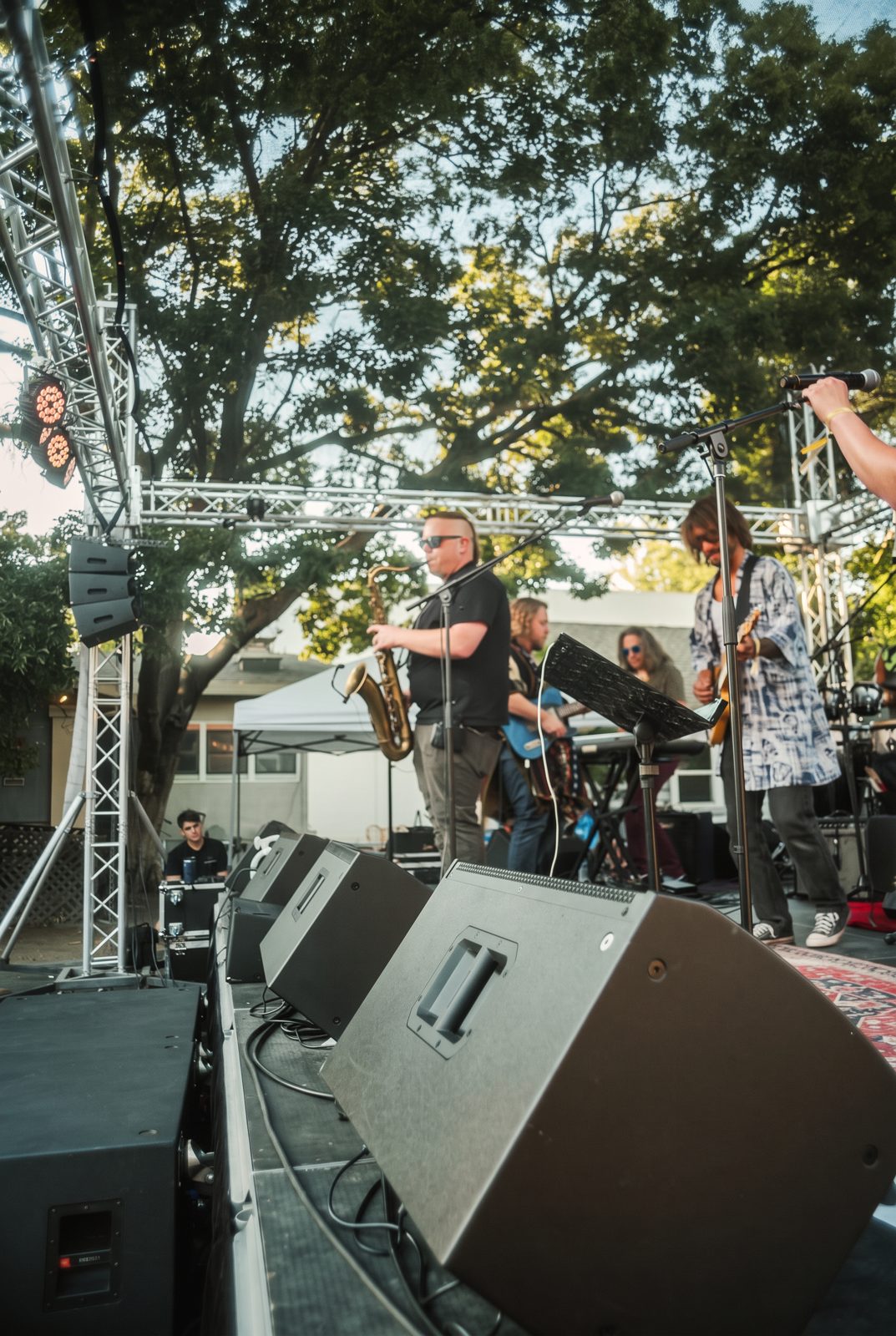 Live band performing on an outdoor stage with monitor wedges and trussing under trees at a Davis daytime concert